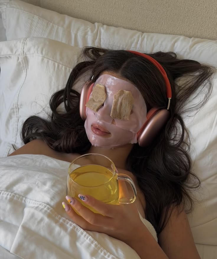 Woman relaxing in bed with headphones, a face mask and a glass mug of golden tea