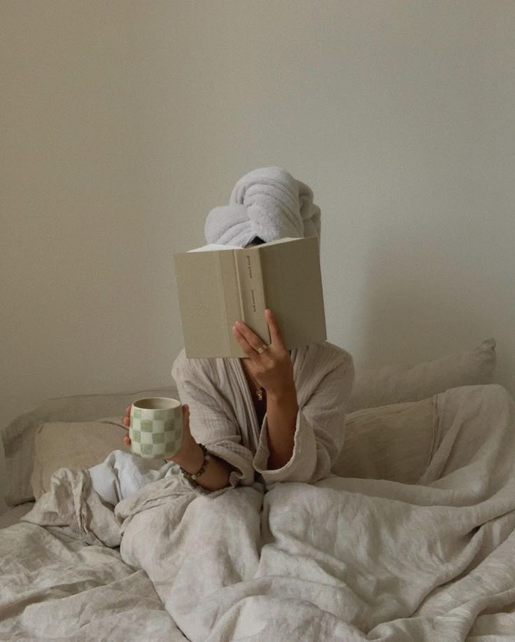 Woman reading a book in bed with a towel wrapped around her hair and a green checkered mug of tea
