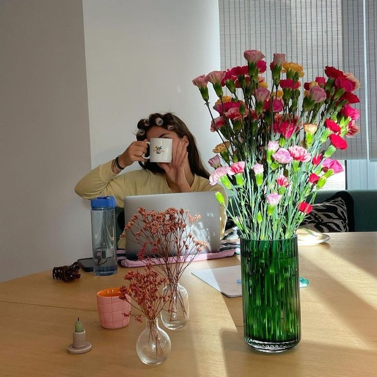 Woman sipping tea from a mug at a sunny table with carnations and dried flowers