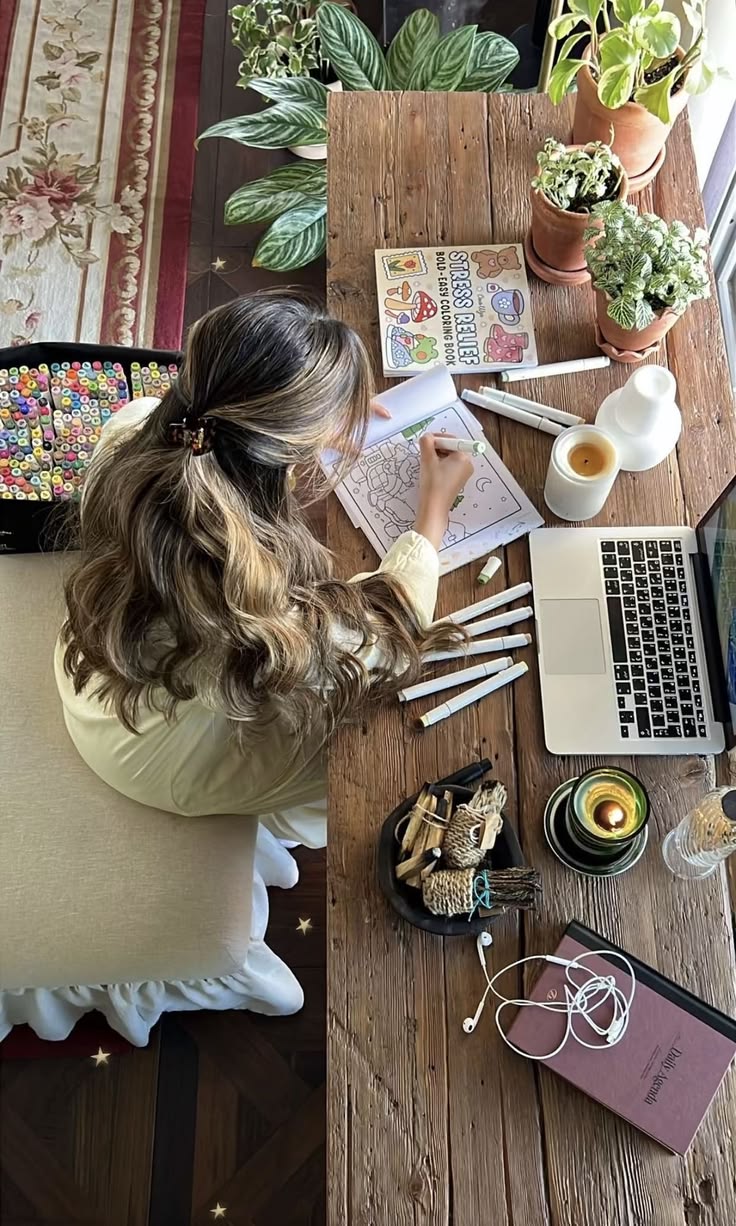 Woman coloring at a wooden desk with a cup of tea, houseplants and a laptop